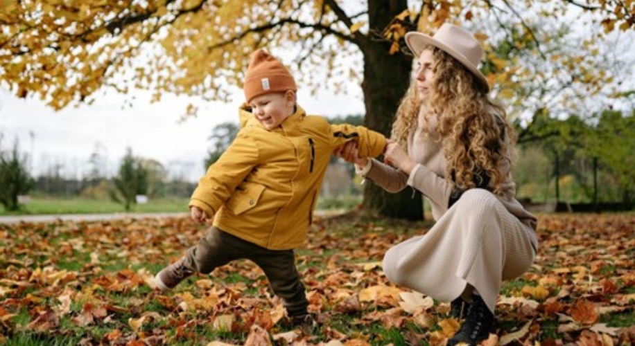 Toddler in a yellow jacket and orange beanie with an adult in a beige outfit amidst autumn leaves.