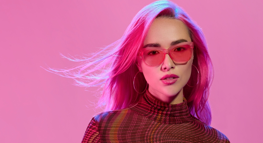 Woman wearing bright pink sunglasses, pink hair, hoop earrings, and a patterned high-neck top.