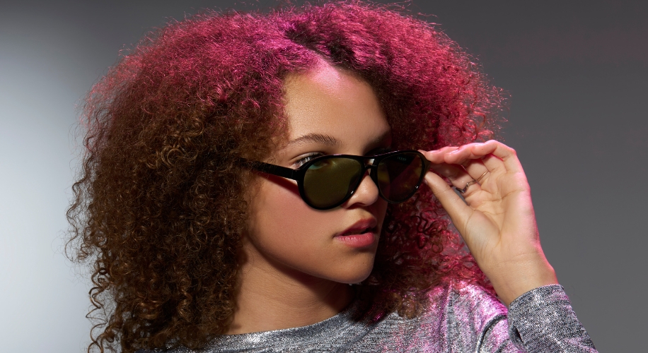 Woman wearing black sunglasses and a shiny silver top, adjusting the sunglasses with her right hand.