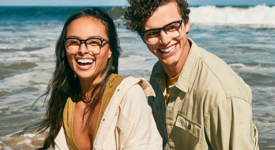 Two people wearing fashionable eyeglasses, smiling at the beach with waves in the background.