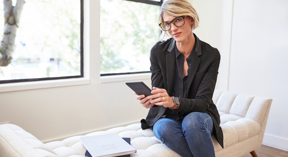 Woman in glasses holding e-reader, seated on beige tufted bench; document nearby says &quot;STYLE INNOVATOR&quot;.