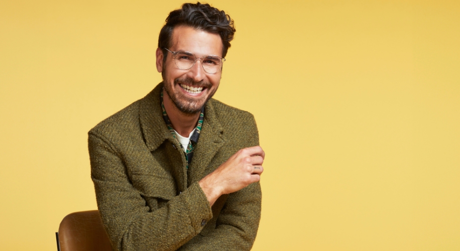 Man in green tweed jacket smiling against yellow background.