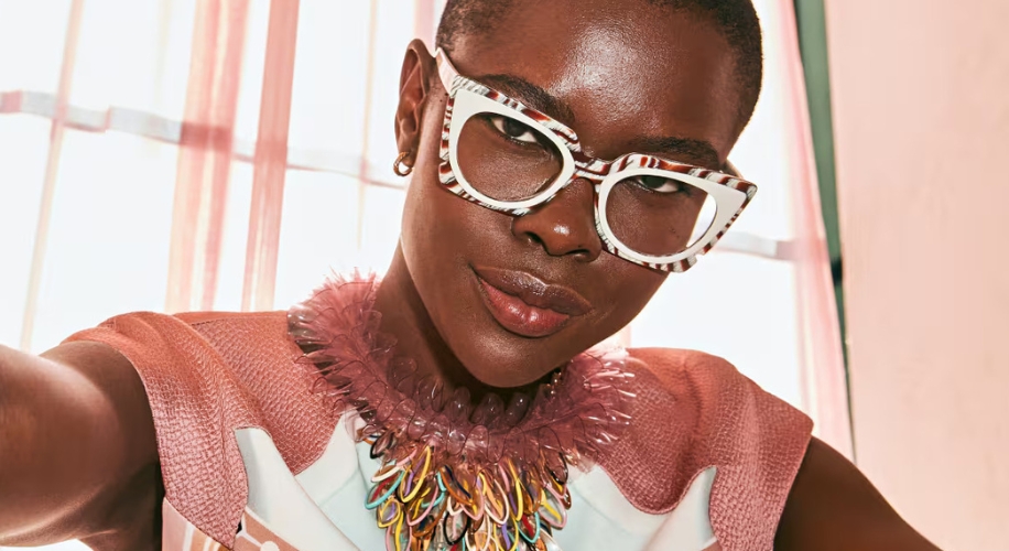 Woman wearing white and black patterned eyeglasses, a textured pink top, and a multicolored statement necklace.