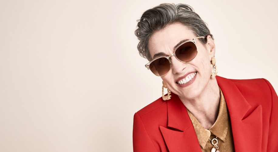 Smiling person wearing oversized sunglasses, gold earrings, red blazer, and brown button-up shirt.