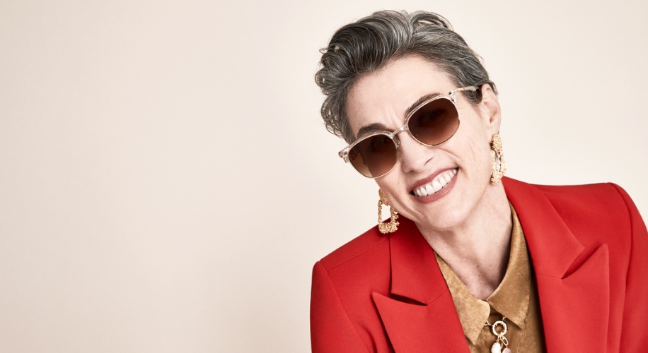 Woman wearing oversized sunglasses, gold earrings, and a red blazer smiles.