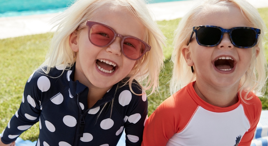 Two smiling children wearing sunglasses and swimwear. One in a polka dot swimsuit and the other in a red and white swimsuit.