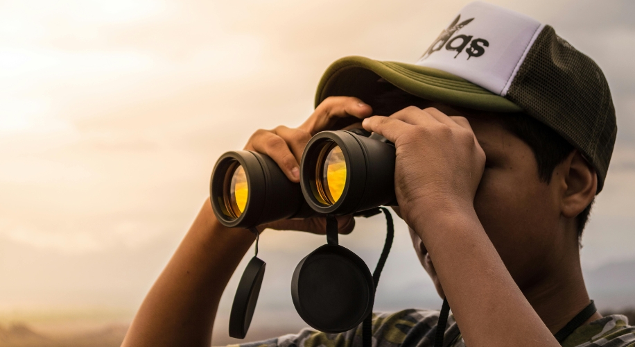 Person using binoculars with yellow tinted lenses, wearing a camo hat with &quot;Ap$&quot; text.