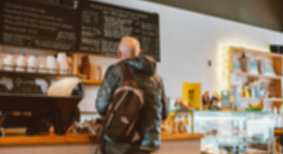 Person with a backpack at a coffee shop counter, with a list of coffee options visible on the chalkboard.