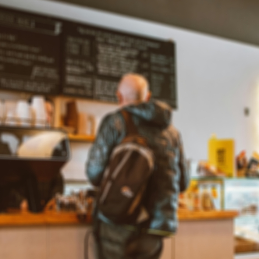 Blurry café scene with a man wearing a hooded jacket and backpack waiting at the counter. Wall menu text not readable.