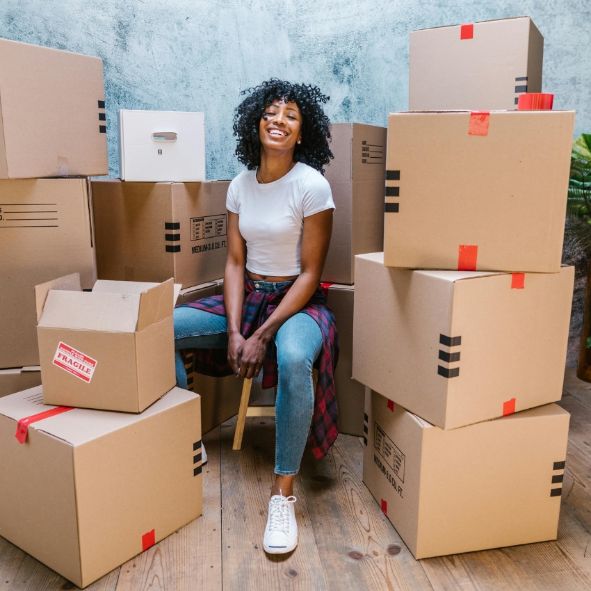 Woman sitting on a stool, surrounded by stacked cardboard boxes; one box labeled "FRAGILE."