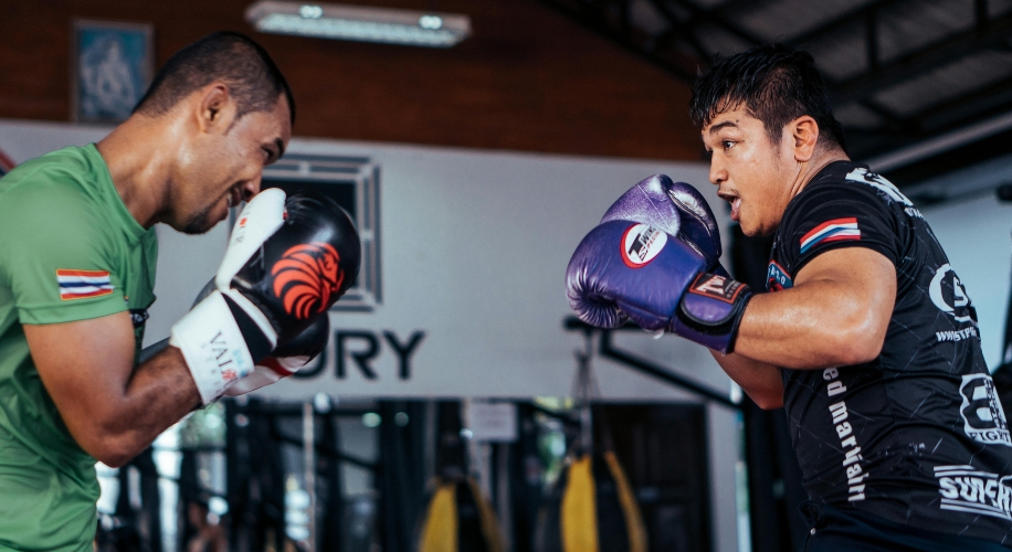 Two fighters spar wearing boxing gloves, one with white gloves and the other with purple gloves.