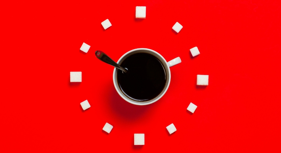 A cup of black coffee with a spoon, surrounded by sugar cubes arranged in a circular pattern on a red background.