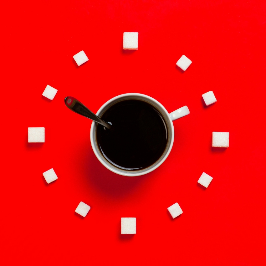 Cup of black coffee with a spoon surrounded by sugar cubes on a red background.