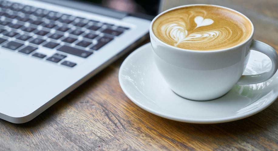 Cup of latte with heart-shaped foam latte art, next to a laptop on a wooden surface.