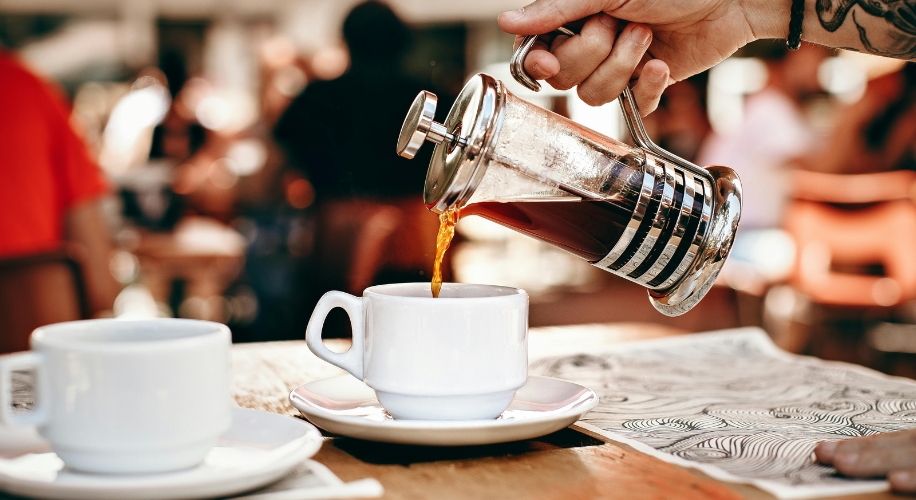 French press coffee being poured into white cup on a wooden table.