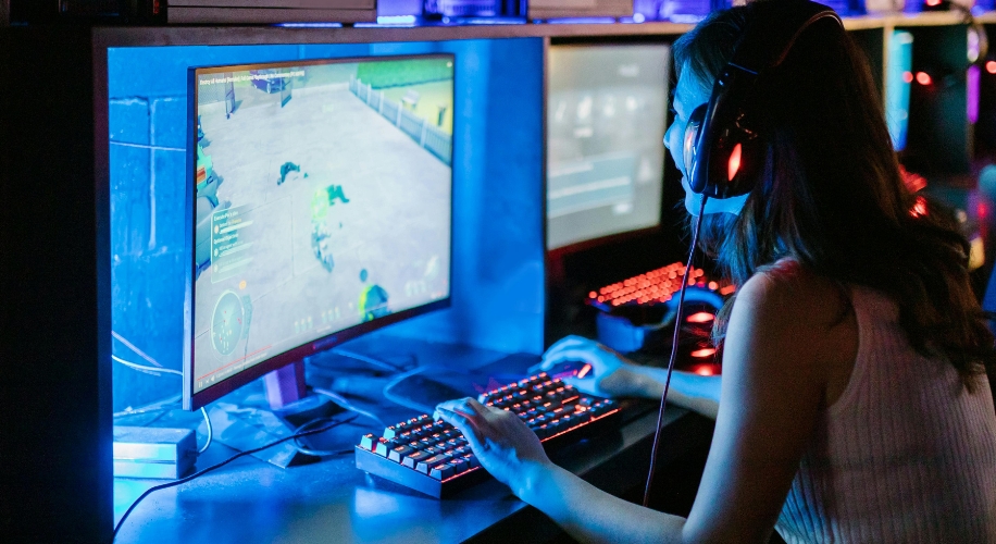 Person playing a game on a curved gaming monitor with a backlit keyboard and wearing headphones.