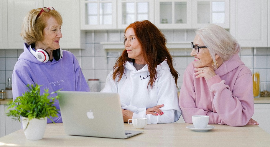 Three women wearing hoodies, one purple with text &quot;RESET OF LIFE WORK!&quot;, one white with unreadable text, and one pink, sit at a table with a laptop and coffee cups.