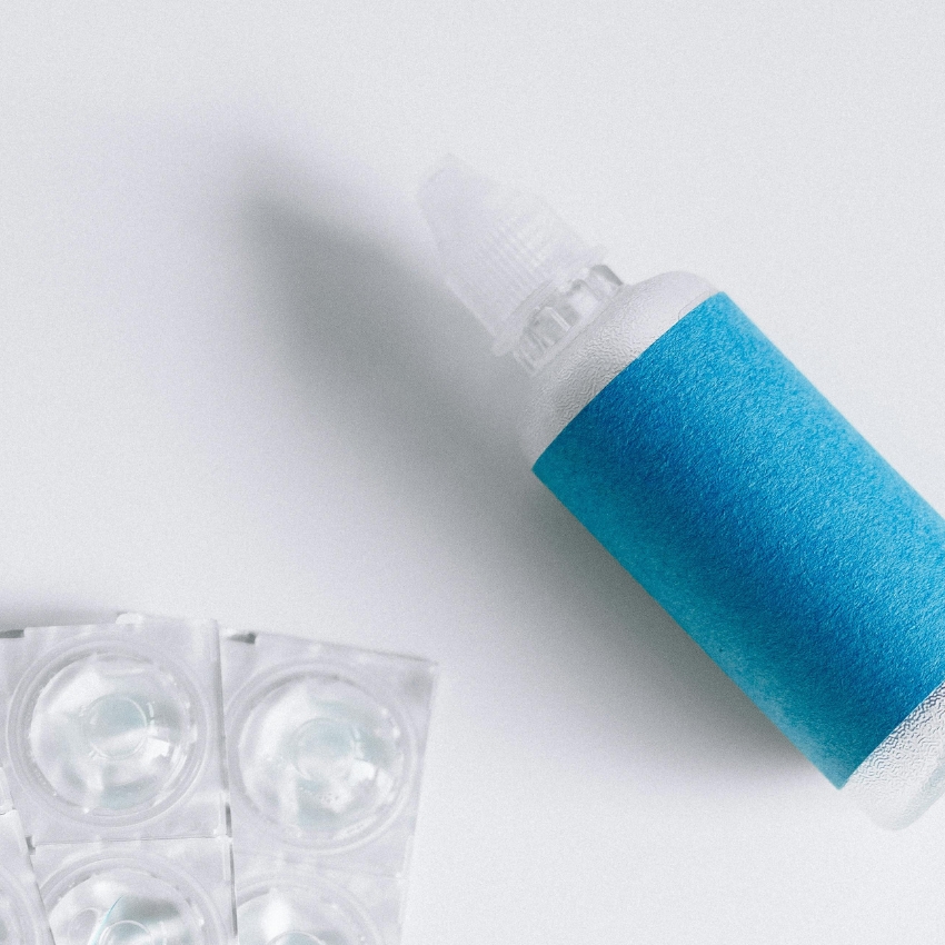 Small plastic eye drop bottle with a blue label beside blister packs of contact lenses.