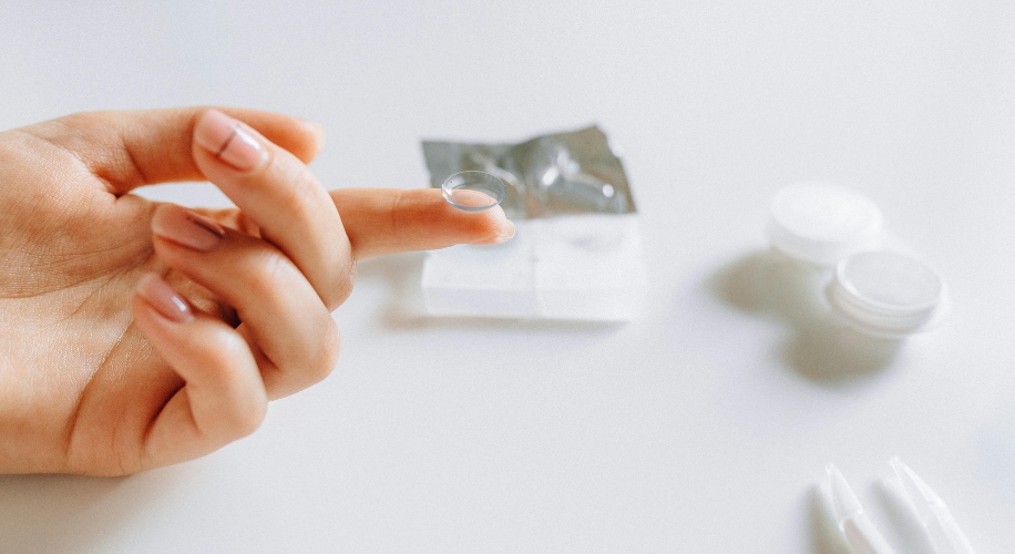 Contact lens balanced on fingertip with lens case and packaging in the background.