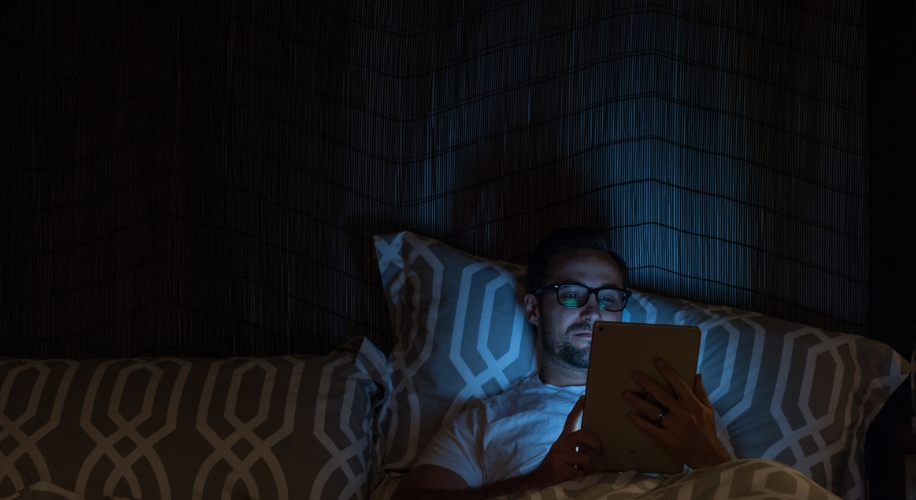 Person wearing glasses using a tablet in bed with patterned pillows and sheets under dim light.