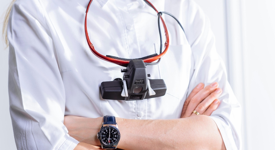 Wearable device hanging on neck strap. Person folding arms, wearing a wristwatch. Keeler logo on device.