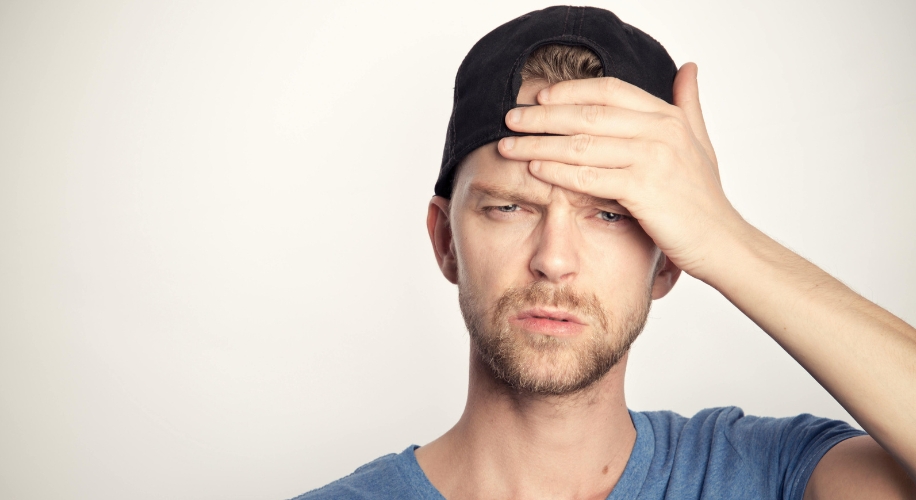 Man in a blue shirt and black cap holding his forehead with a concerned expression.