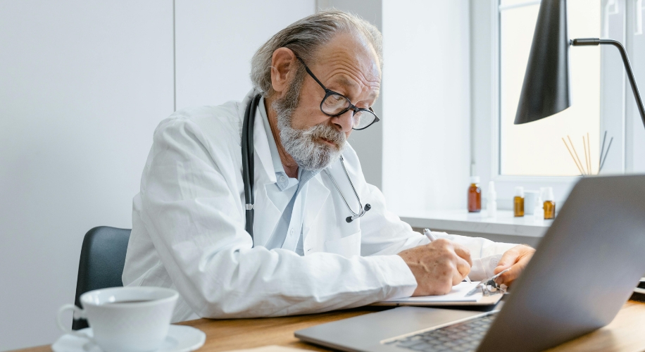 Elderly doctor wearing a stethoscope, writing on paper at a desk with a laptop and coffee cup.