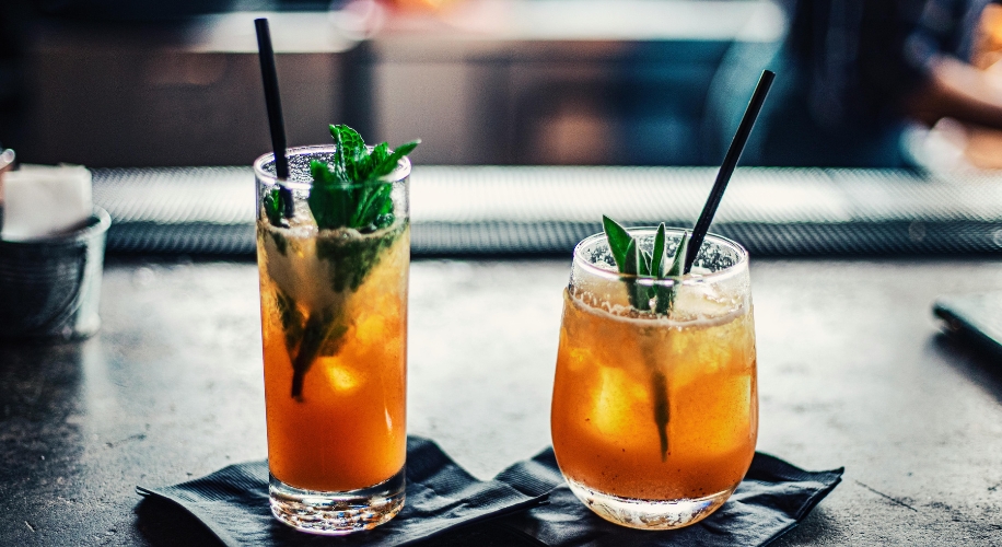 Two orange cocktails with black straws, garnished with fresh green leaves, on dark napkins.