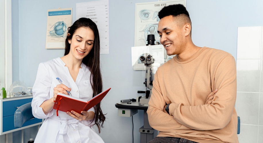 Optometrist writing in a red notebook, seated with a smiling patient in an eye examination room.