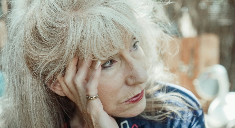 Elderly woman with long grey hair resting her head on her hand, wearing a gold ring and blue clothing.