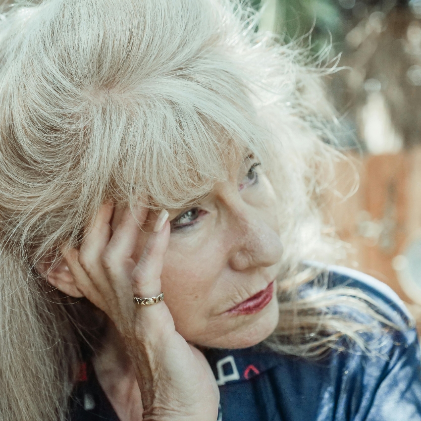 Woman with long grey hair wearing a gold ring, resting her head on her hand.