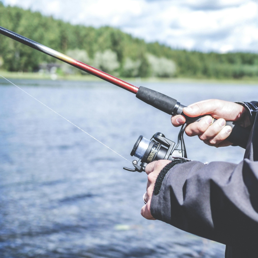 Hands holding a fishing rod and reel by a lakeside.