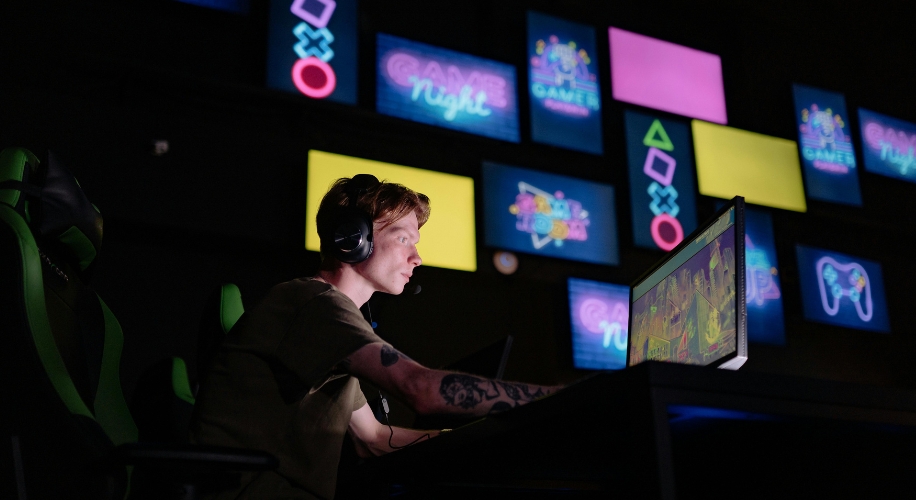 Person gaming on a computer with neon signs in the background displaying &quot;Game Night&quot; and various symbols.