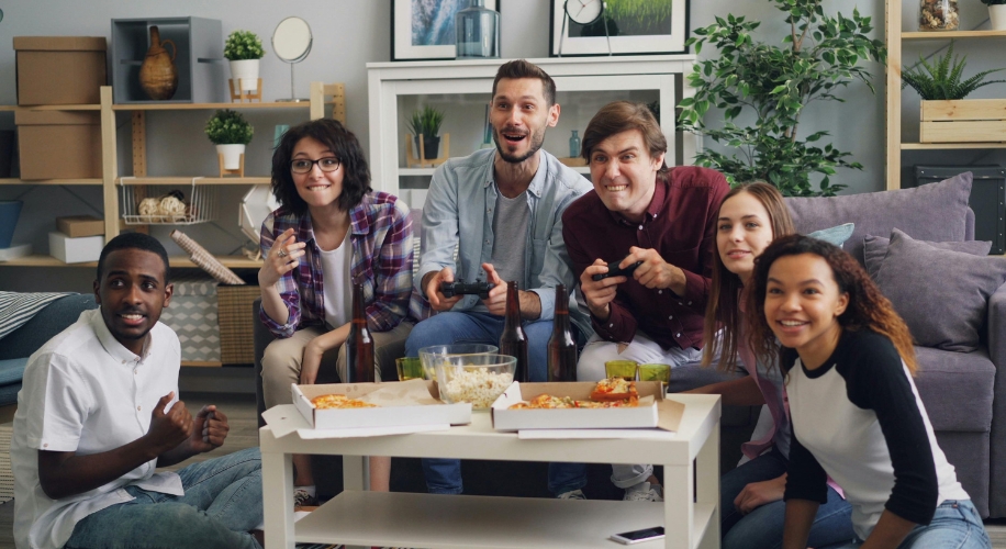 Group of friends playing video games, sitting around a table with pizza, popcorn, and drinks.