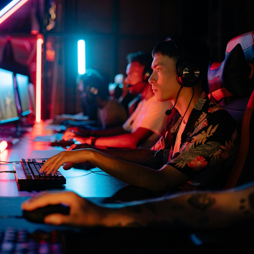 Gamers with headsets intensely playing on computers with illuminated keyboards and colorful lighting.