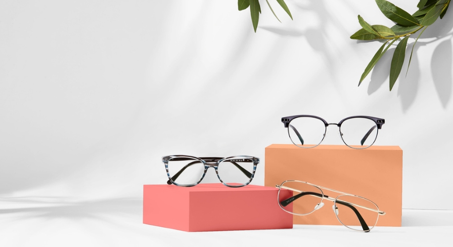 Three pairs of eyeglasses on pink and orange blocks with a white background and green plant leaves.