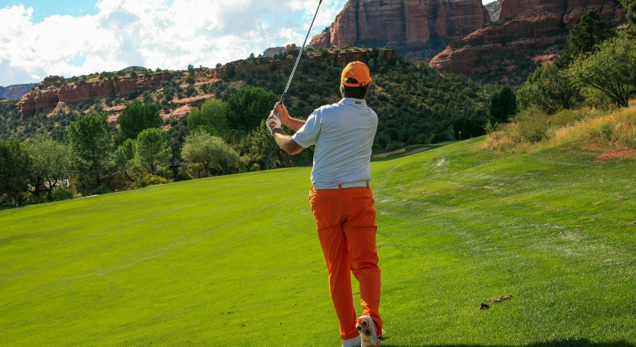 Golfer in orange pants and cap, white shirt, swinging a golf club on a lush green fairway.