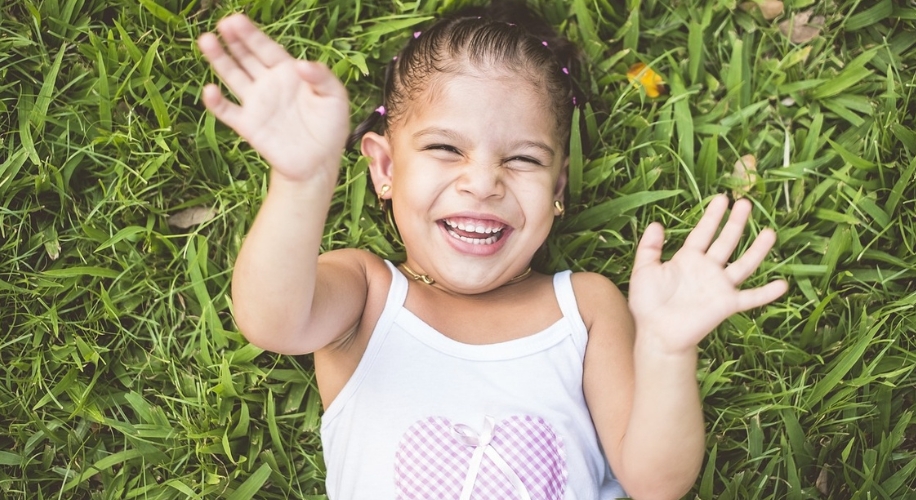 Child smiling with hands up, lying on grass, wearing a white tank top with a pink heart design.