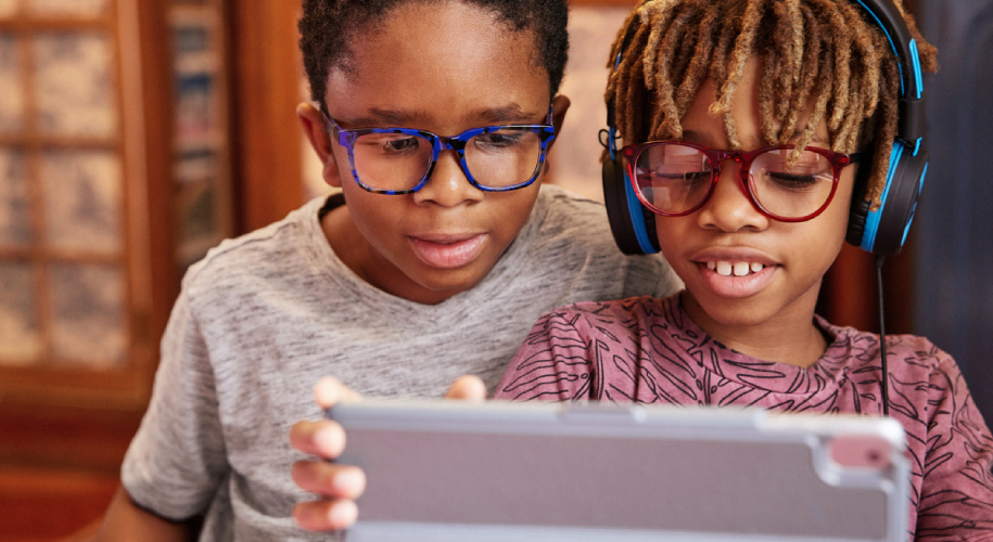 Two children wearing glasses use a tablet; one also wears blue headphones.