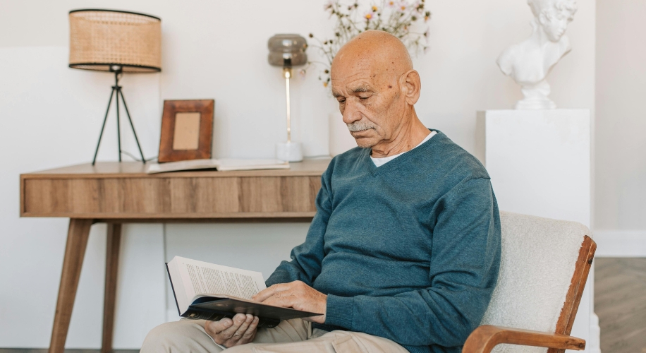 Elderly man wearing a blue sweater, seated on a beige chair, reading a book.