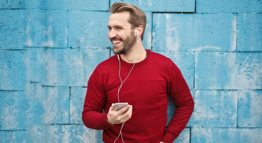 Man wearing earphones and red sweater, holding a smartphone, smiling against a blue wall.