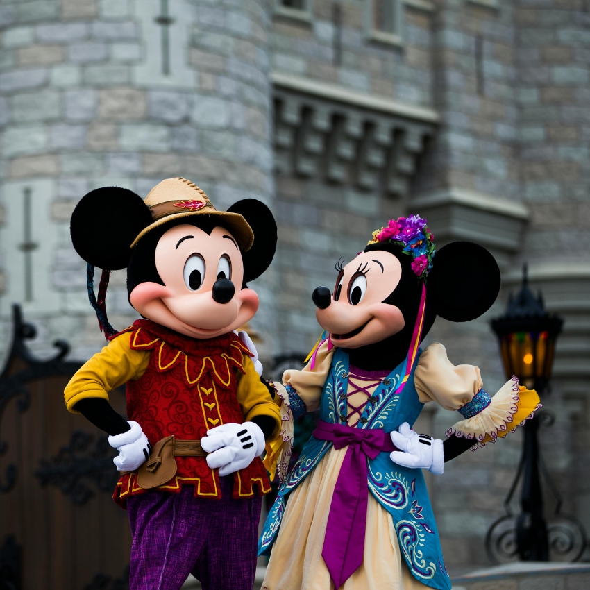 Mickey Mouse and Minnie Mouse dressed in colorful traditional outfits, standing and smiling.