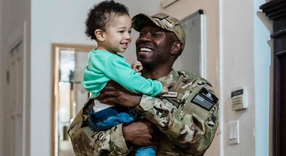 Soldier in camo uniform smiling and holding a child.