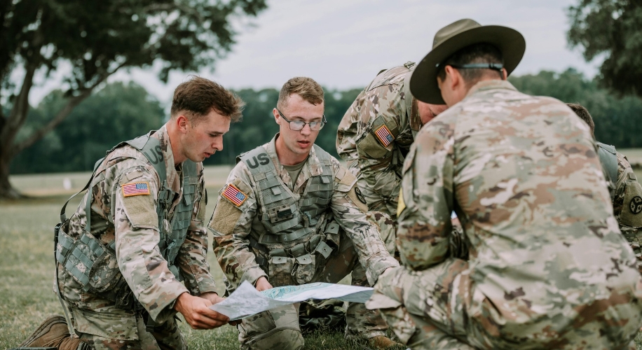 Soldiers in uniform gather in a circle on a grassy field, studying a map together.