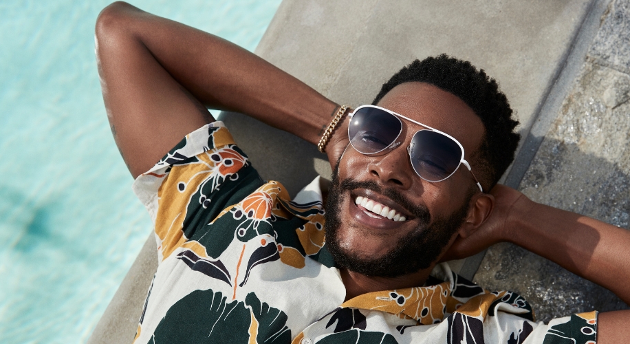 Man wearing a floral shirt and sunglasses, smiling and relaxing by a pool.