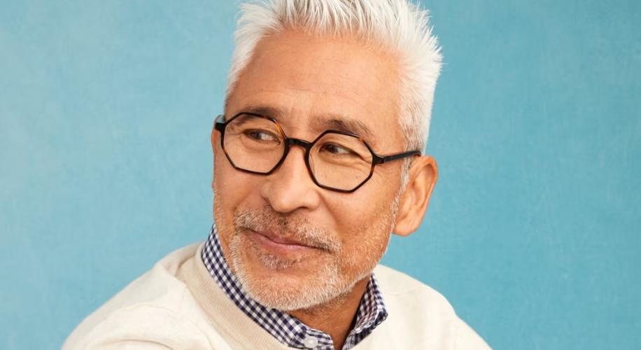 Man wearing black rectangular glasses and a white sweater over a checkered shirt against a blue background.