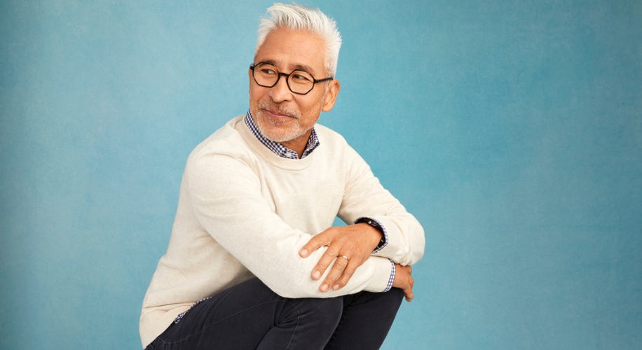 Man wearing a white sweater and glasses, squatting against a blue background.