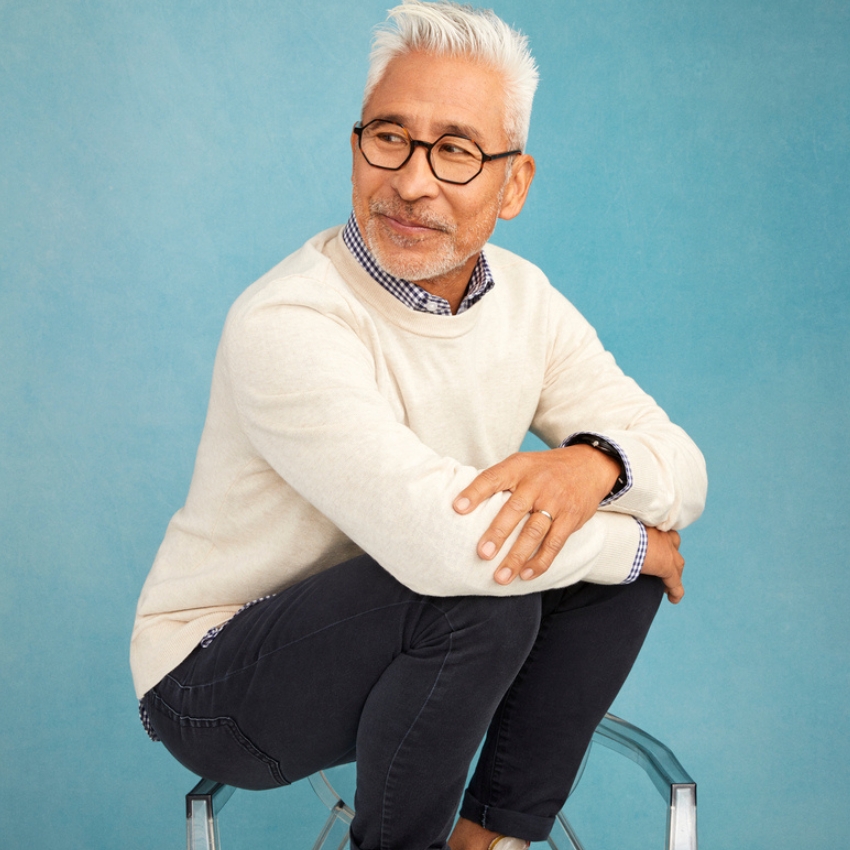 Man wearing a cream-colored sweater with a white and blue checkered shirt, dark-rimmed glasses, and black pants.