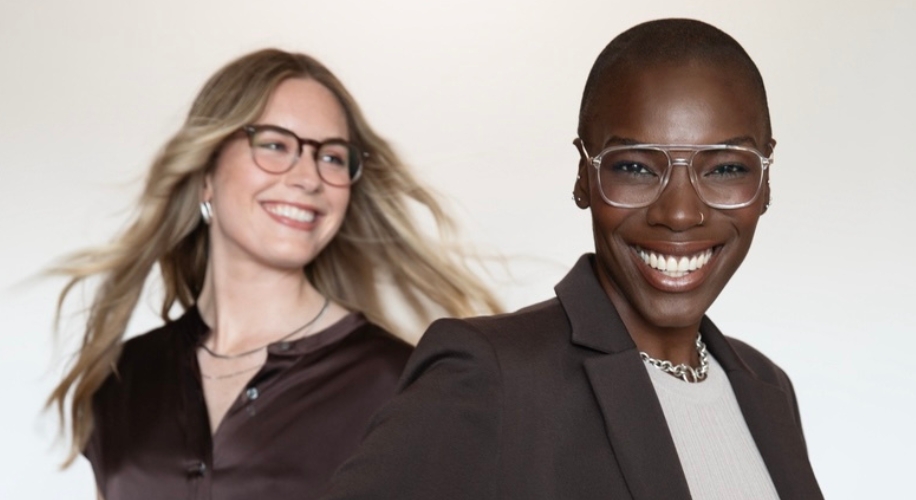 Two women smiling, both wearing stylish eyeglasses; one with a brown blouse and the other with a blazer.