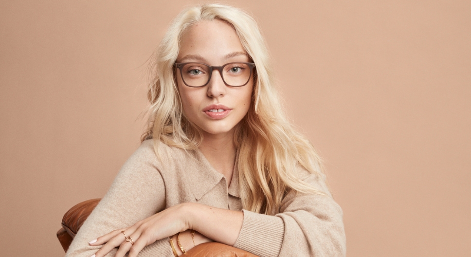 Woman wearing brown-rimmed glasses, light tan sweater, blonde hair, and gold bracelet on left wrist.